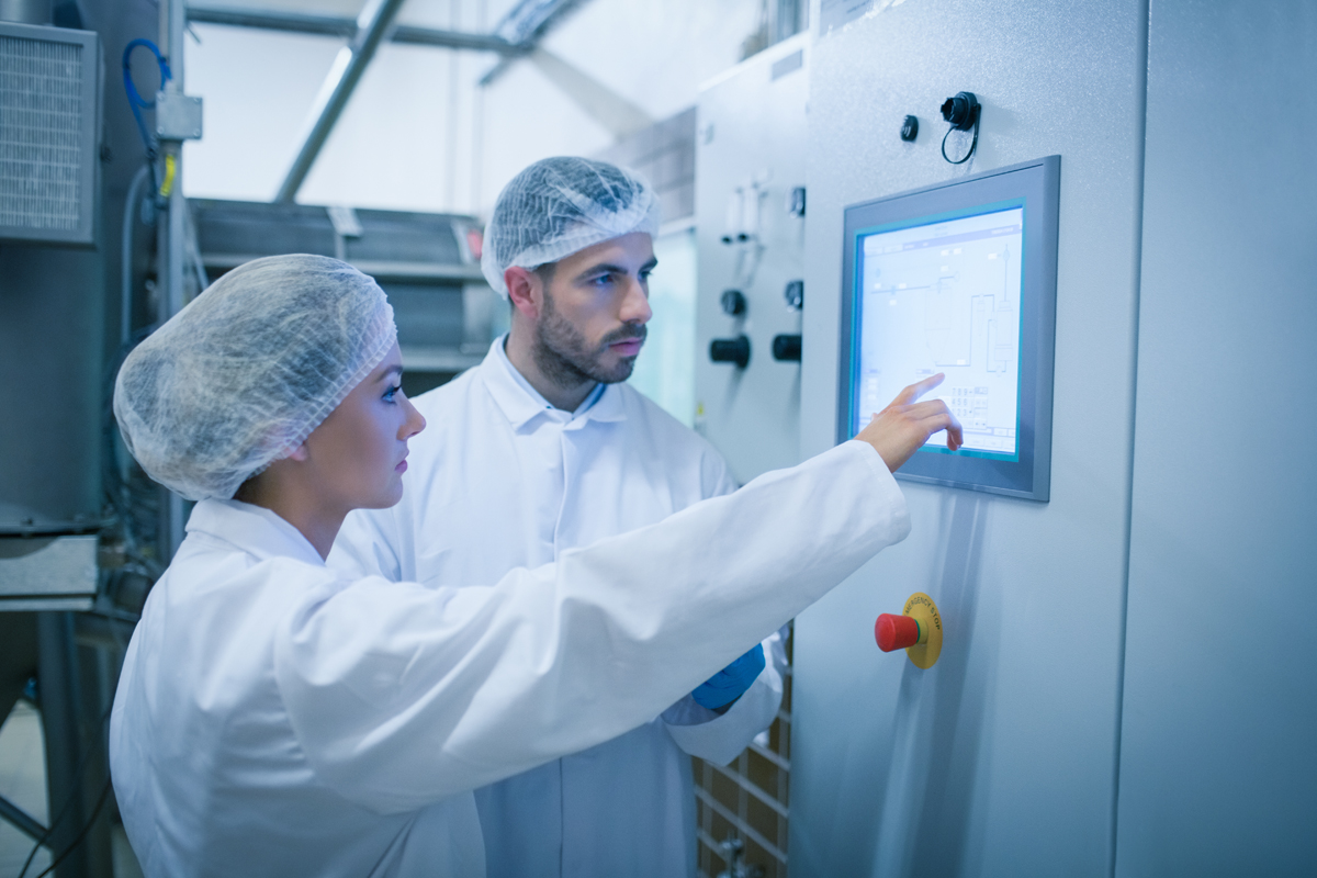 Two food science technicians in white coats and hairnets operate a computerized control panel.